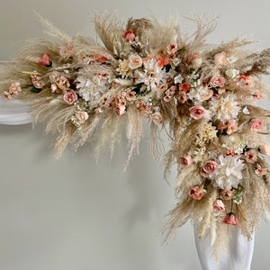 May include: A floral arch arrangement featuring pampas grass, roses, and dahlias in shades of cream, peach, and blush. The arrangement is set against a white backdrop, creating a soft and romantic aesthetic. Ideal for weddings and special events.
