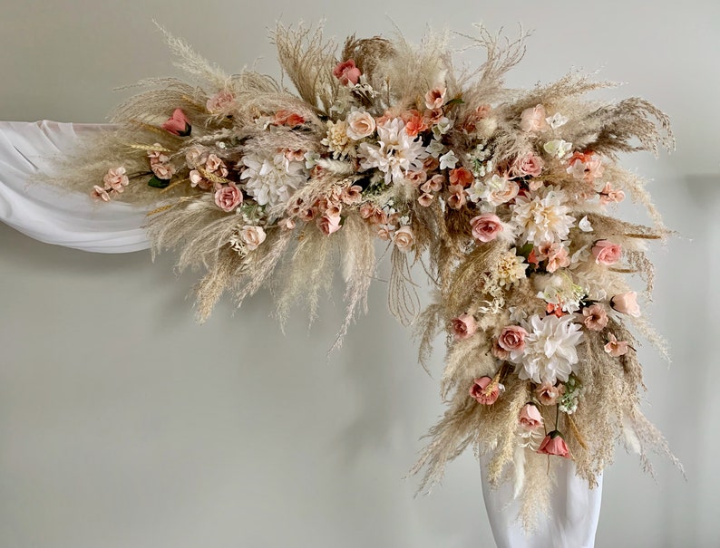 May include: A floral arch arrangement featuring pampas grass, roses, and dahlias in shades of cream, peach, and blush. The arrangement is set against a white backdrop, creating a soft and romantic aesthetic. Ideal for weddings and special events.
