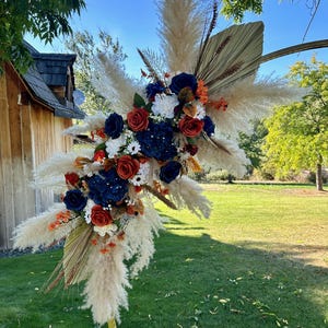 May include: A decorative floral arrangement with navy blue, red, and white flowers, complemented by dried palm leaves and pampas grass. The arrangement is set against a backdrop of green grass and a clear blue sky, suitable for weddings or special events.