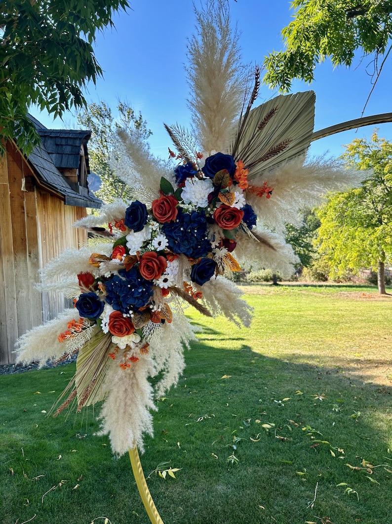 May include: A decorative floral arrangement with navy blue, red, and white flowers, complemented by dried palm leaves and pampas grass. The arrangement is set against a backdrop of green grass and a clear blue sky, suitable for weddings or special events.
