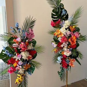以下が含まれることがあります： 2つの背の高い金のスタンドに、熱帯の花の飾り付けが施されています。飾り付けには、蘭、アジサイ、ユリなど、さまざまな色の花と緑の葉が使われています。