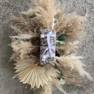 May include: A rectangular gift wrapped in patterned brown paper and tied with white ribbon, centered on a display of dried pampas grass, a fan-shaped dried palm leaf, and white flowers. The arrangement is set against a gray background.