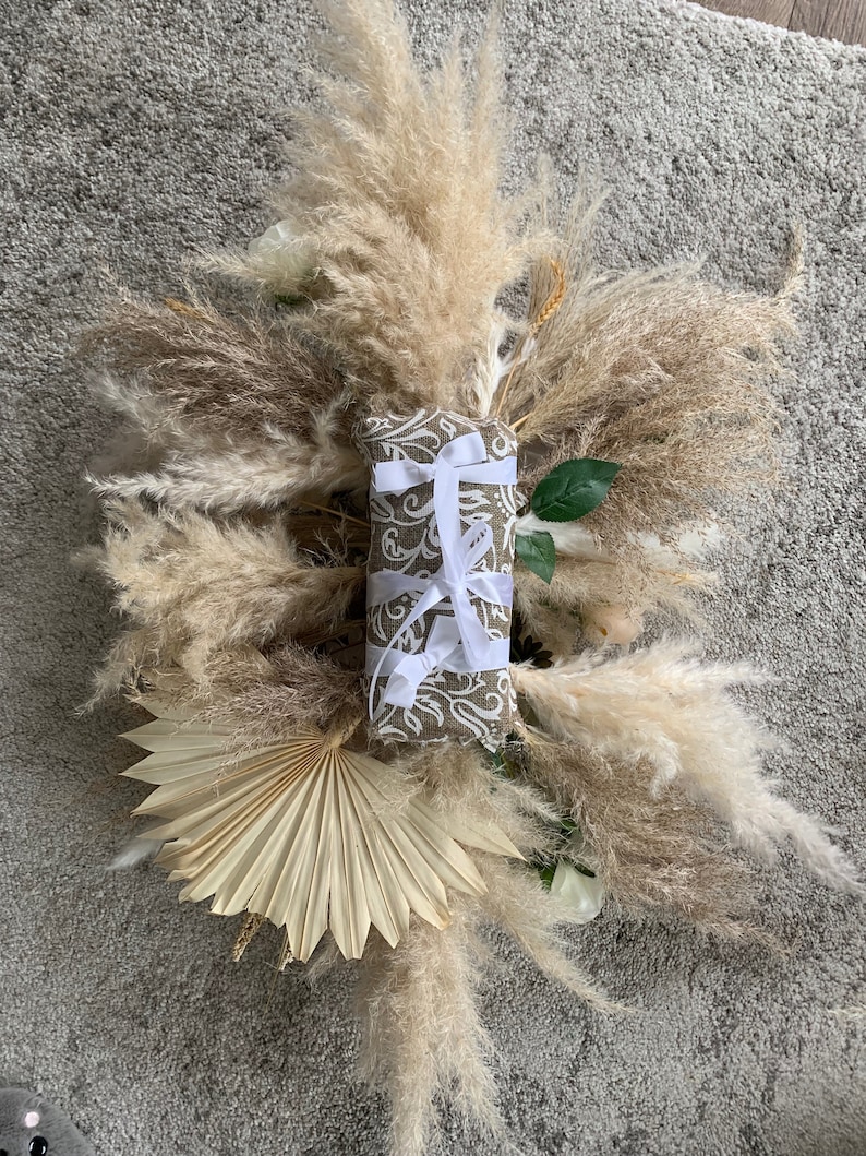 May include: A rectangular gift wrapped in patterned brown paper and tied with white ribbon, centered on a display of dried pampas grass, a fan-shaped dried palm leaf, and white flowers. The arrangement is set against a gray background.