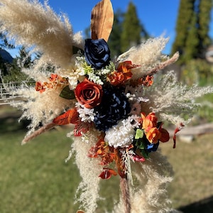 May include: A decorative floral arrangement with pampas grass, navy blue, orange, and red flowers. The arrangement includes a variety of textures and colours, with a wooden accent. The backdrop is a clear blue sky and green grass.