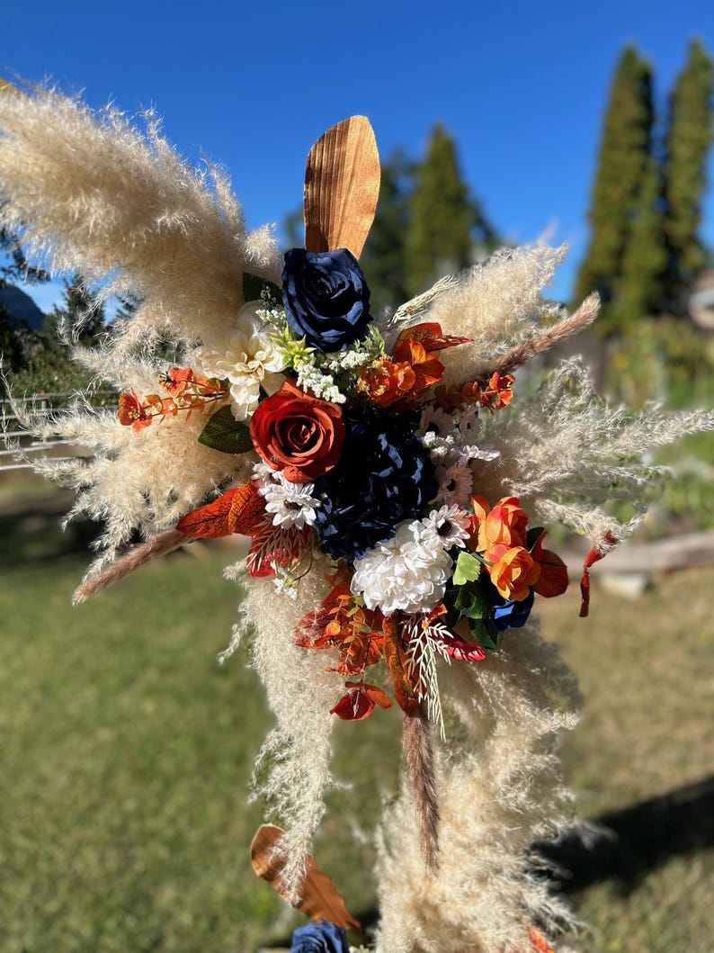 May include: A decorative floral arrangement with pampas grass, navy blue, orange, and red flowers. The arrangement includes a variety of textures and colours, with a wooden accent. The backdrop is a clear blue sky and green grass.