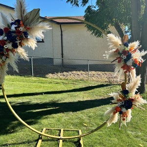 May include: A circular golden metal wedding arch adorned with floral displays. The arch is positioned on a grassy lawn. The floral arrangements feature pampas grass, dried palm leaves, and flowers in red, white, and blue hues.
