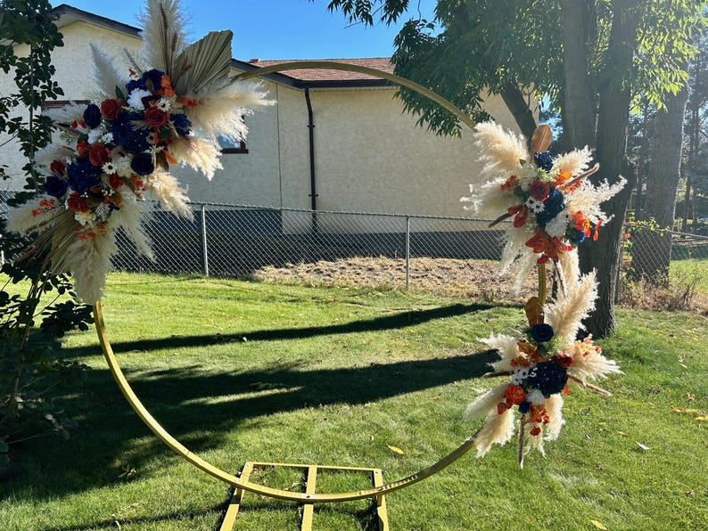 May include: A circular golden metal wedding arch adorned with floral displays. The arch is positioned on a grassy lawn. The floral arrangements feature pampas grass, dried palm leaves, and flowers in red, white, and blue hues.