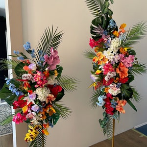 以下が含まれることがあります： 2つの高い金色の金属製のスタンドに、熱帯の花の飾り付けが施されています。飾り付けには、ピンク、オレンジ、イエロー、レッド、ブルー、ホワイトなど、さまざまな色の花が使われています。花は、一番上の花が最も高く、一番下の花が最も低い、滝のように配置されています。スタンドは木製の床の上に置かれています。