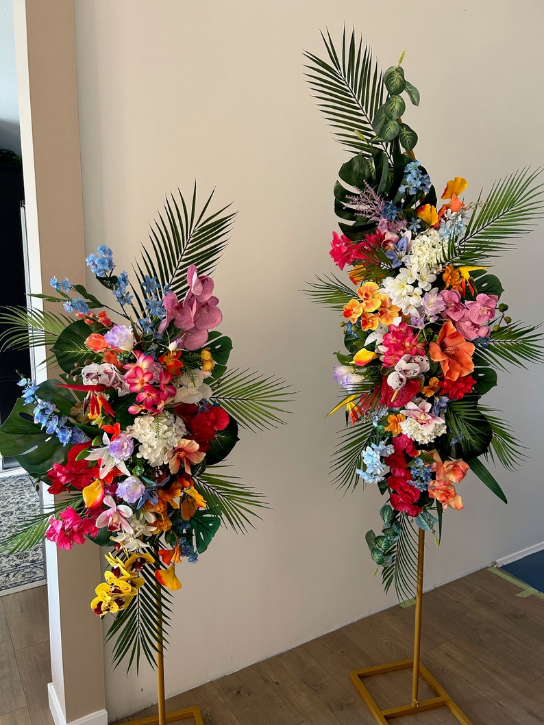 以下が含まれることがあります： 2つの高い金色の金属製のスタンドに、熱帯の花の飾り付けが施されています。飾り付けには、ピンク、オレンジ、イエロー、レッド、ブルー、ホワイトなど、さまざまな色の花が使われています。花は、一番上の花が最も高く、一番下の花が最も低い、滝のように配置されています。スタンドは木製の床の上に置かれています。