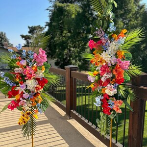 以下が含まれることがあります： 2つの高い金色の金属製のスタンドに、熱帯の花の飾り付けが施されています。スタンドは、黒い金属製の柵のある木製のデッキの上に置かれています。花は、ピンク、赤、オレンジ、黄色、白、青のミックスです。