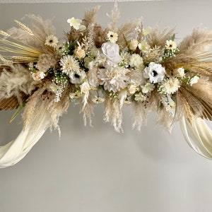 May include: A white and beige floral arrangement with pampas grass and palm leaves, draped over a birch wood archway with white fabric.