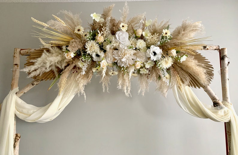May include: A white and beige floral arrangement with pampas grass and palm leaves, draped over a birch wood archway with white fabric.