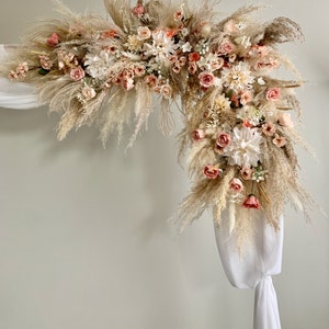 May include: An archway decorated with dried pampas grass, white and peach-colored flowers, and white fabric draping. The floral arrangement is dense and full, creating a natural and elegant aesthetic. Ideal for weddings or special events.