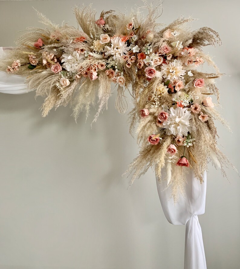 May include: An archway decorated with dried pampas grass, white and peach-colored flowers, and white fabric draping. The floral arrangement is dense and full, creating a natural and elegant aesthetic. Ideal for weddings or special events.