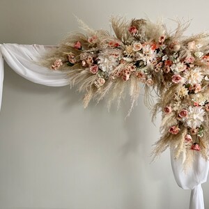 May include: A decorative wedding arch featuring a floral arrangement. The arch is adorned with a mix of pampas grass, roses, and other flowers in shades of cream, peach, and pink. White fabric drapes from the wooden frame.