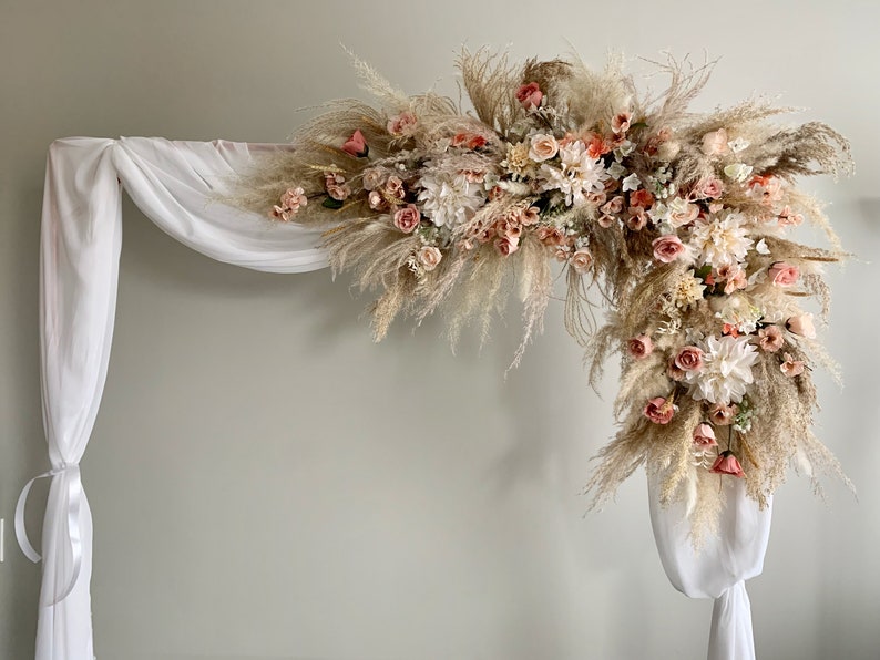May include: A decorative wedding arch featuring a floral arrangement. The arch is adorned with a mix of pampas grass, roses, and other flowers in shades of cream, peach, and pink. White fabric drapes from the wooden frame.