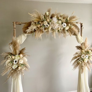 May include: A rustic wooden archway decorated with dried pampas grass, white flowers, and greenery. The archway is adorned with white fabric drapes.