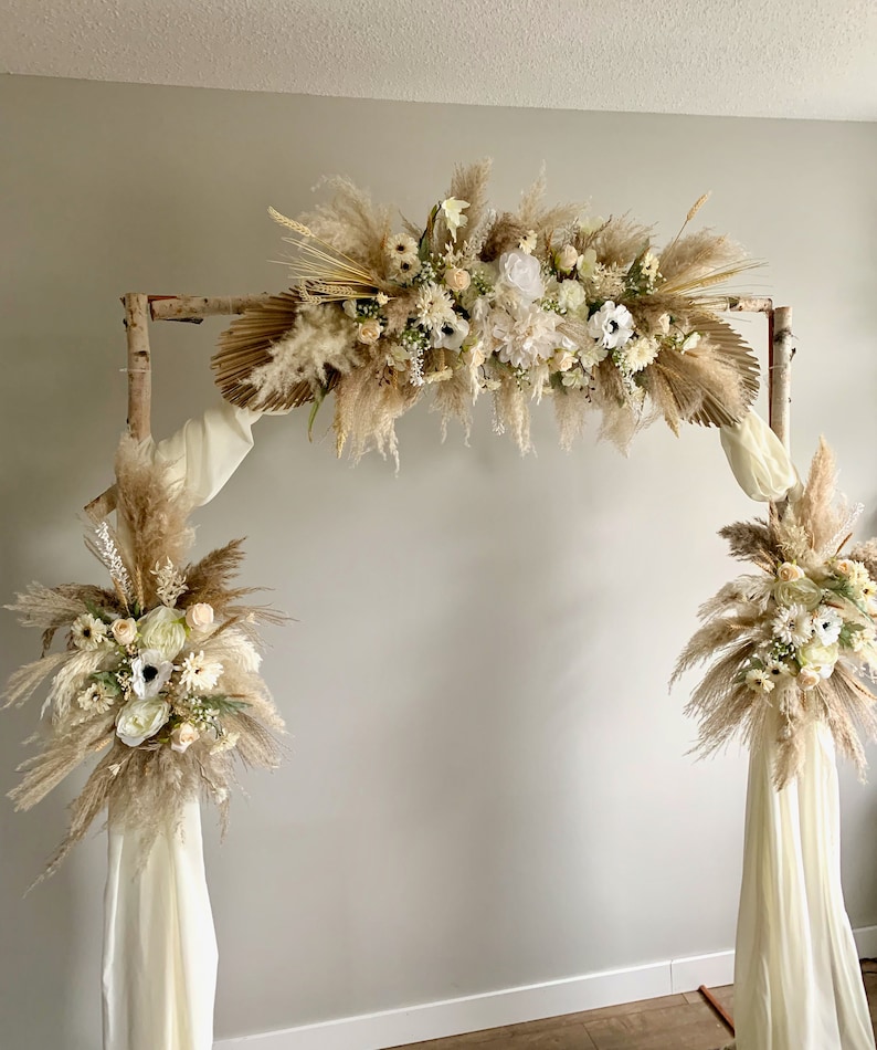May include: A rustic wooden archway decorated with dried pampas grass, white flowers, and greenery. The archway is adorned with white fabric drapes.