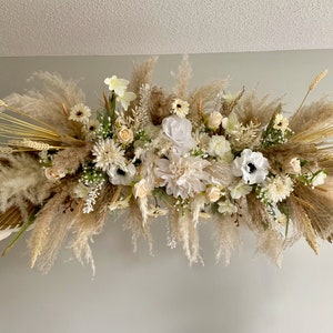 May include: A white and cream floral arrangement with pampas grass and dried wheat. The arrangement is hanging from a wooden archway.