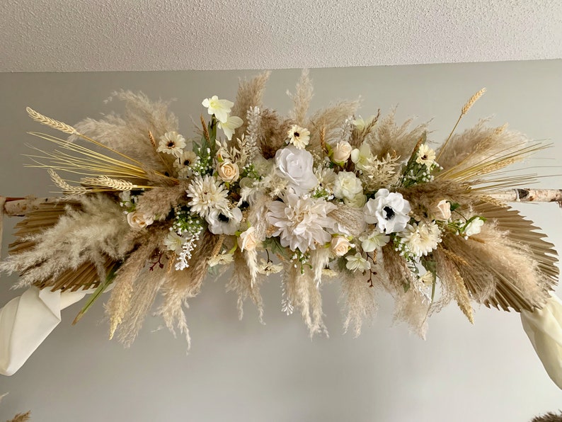 May include: A white and cream floral arrangement with pampas grass and dried wheat. The arrangement is hanging from a wooden archway.