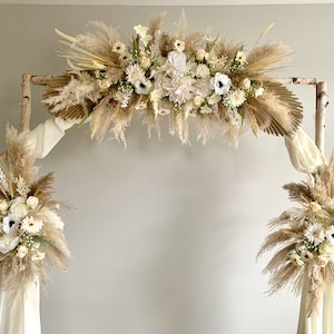 May include: A rustic wedding arch made of birch branches decorated with dried pampas grass, white flowers, and greenery.