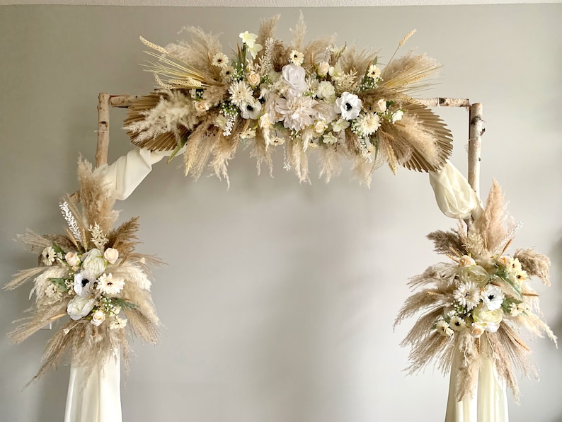 May include: A rustic wedding arch made of birch branches decorated with dried pampas grass, white flowers, and greenery.