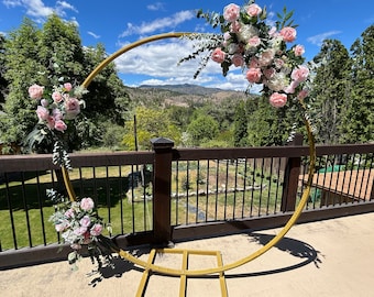 May include: A gold metal archway decorated with pink and white flowers, greenery, and eucalyptus leaves. The archway is set up outdoors on a wooden deck with a view of a mountain range in the background.