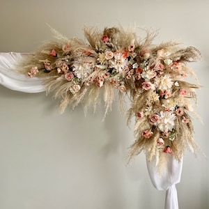 May include: A decorative floral arch featuring a mix of dried pampas grass, blush roses, and white dahlias. The arrangement is accented with delicate greenery and draped with white fabric, creating a soft, romantic aesthetic, perfect for weddings or events.