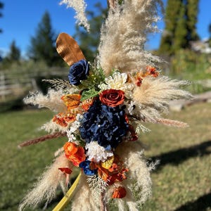 May include: A decorative floral arrangement with pampas grass, navy blue, orange, and red flowers. The arrangement includes a variety of textures and colours, with a focus on natural elements. Suitable for weddings or special events.