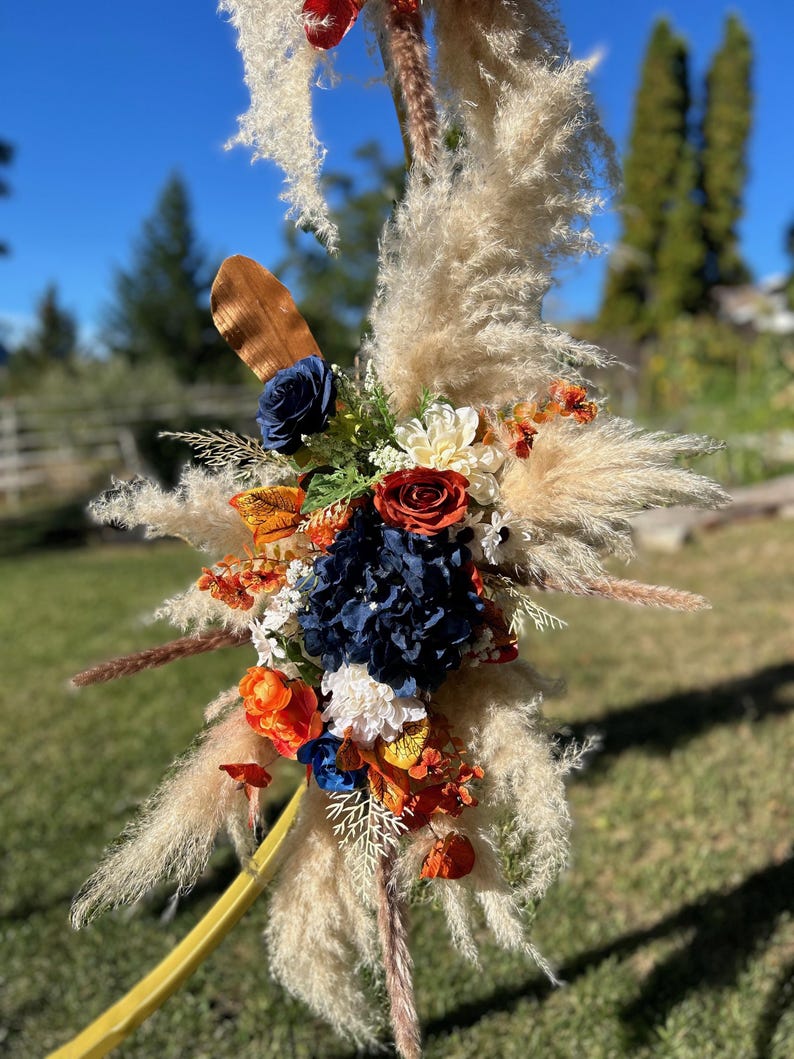 May include: A decorative floral arrangement with pampas grass, navy blue, orange, and red flowers. The arrangement includes a variety of textures and colours, with a focus on natural elements. Suitable for weddings or special events.