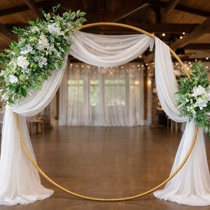 May include: A gold metal wedding arch with white fabric draping and floral arrangements. The arch is circular and stands in a large room with tables and chairs in the background. The floral arrangements include white flowers and greenery.