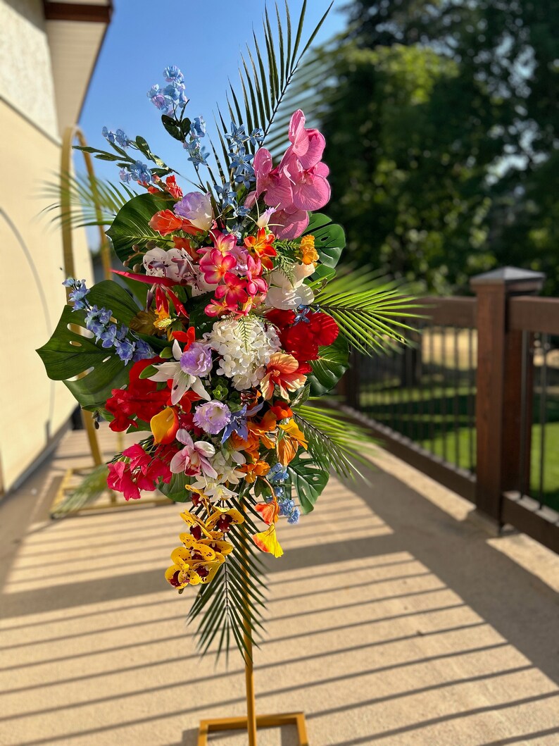 以下が含まれることがあります： ピンク、赤、黄、オレンジ、青、白の花を使ったトロピカルなフラワーアレンジメント。アレンジメントは金色の金属製のスタンドに置かれ、ヤシの葉が含まれています。