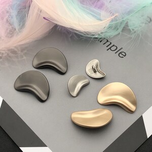 May include: A selection of crescent-shaped metal buttons in gunmetal, silver, and gold. The buttons have a smooth, curved design and are displayed on a grey surface. Some buttons have a shank on the back for sewing.