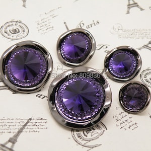 May include: Four purple rhinestone buttons with a silver metal rim. The buttons are of different sizes.