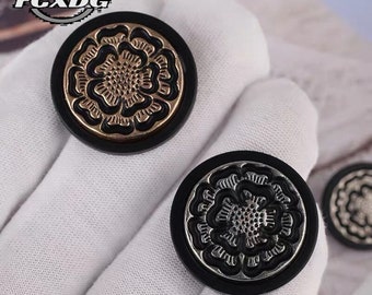 May include: Two black round buttons with a gold and silver floral design. The buttons have a raised design and are surrounded by a black border.