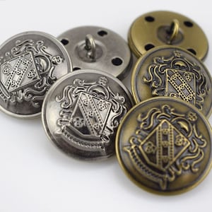 Puede incluir: Un conjunto de cinco botones decorativos con un diseño de escudo de armas. Los botones son de metal y tienen un acabado plateado y bronce.