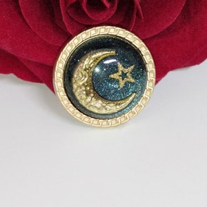 May include: A gold-toned button with a crescent moon and star design. The moon is gold with a detailed design and the star is gold with five points. The background is a dark teal with glitter.