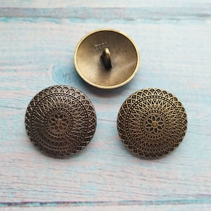 以下が含まれることがあります： マンダラの装飾的なデザインが施された、丸いブロンズ色のボタンが3つあります。ボタンの裏面には、"1983"という数字が浮き彫りにされています。