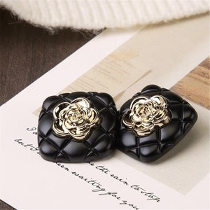 May include: A pair of black square-shaped earrings with a gold rose design. The earrings have a quilted pattern.
