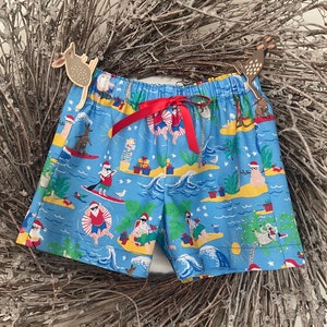 May include: Blue and white patterned shorts with a red ribbon tie. The pattern features Santa Claus characters on a beach with palm trees, surfboards, and presents.
