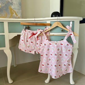 May include: A pink and white striped pajama set, including a tank top and shorts, hangs on wooden hangers. The fabric features a repeating pattern of cherries and floral designs. The tank top has tie straps, and the shorts have a red ribbon tie.