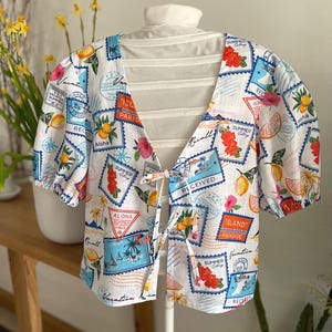 May include: White linen blouse with a tie front and short sleeves. The blouse has a colorful print of stamps with vacation-themed designs, including lemons, palm trees, and the words "Aloha", "Island Paradise", and "Vacation Vibes".