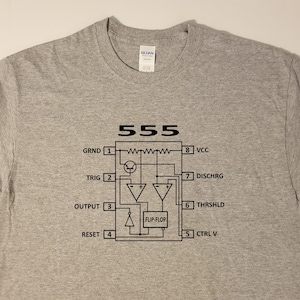 May include: A gray t-shirt with a black graphic of a 555 timer circuit diagram. The diagram includes the pin numbers and labels for each pin, including GND, TRIG, OUTPUT, RESET, VCC, DISCHRG, THRSHLD, and CTRL V.
