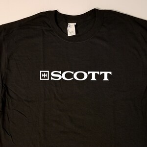 Puede incluir: Camiseta negra con un logotipo blanco que dice "SCOTT" con un cuadrado y tres l&iacute;neas verticales a la izquierda de la palabra.