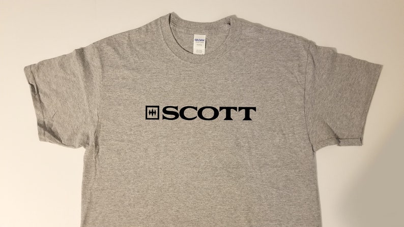 Puede incluir: Una camiseta gris con un logotipo negro que dice "SCOTT" con un cuadrado que contiene una "H" estilizada a la izquierda de la palabra.