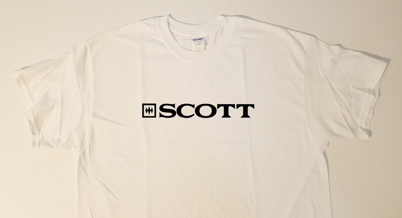 Puede incluir: Camiseta blanca con un logotipo negro que dice "SCOTT" con un peque&ntilde;o cuadrado negro que contiene tres l&iacute;neas horizontales a la izquierda de la palabra.
