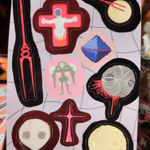 May include: A sheet of stickers featuring various designs, including a cross with a figure, a stylized eye, and a creature. The stickers have a black outline and are on a light purple background.
