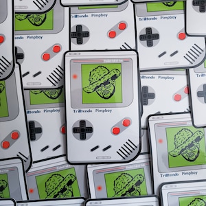 May include: A set of 10 stickers featuring a pixelated cartoon character wearing a hat and sunglasses. The stickers are designed to look like a Game Boy Color game cartridge. The text on the stickers reads "Trilltendo Pimpboy" and "Pocket full of fun".