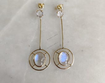 May include: A pair of gold earrings with a round, white moonstone set in a gold cage design. The earrings have a small, white gemstone at the top.