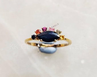 以下が含まれることがあります： マルキーズ型の青い宝石が、より小さな多色の宝石で囲まれた、金の指輪。指輪は白い背景の上に置かれている。